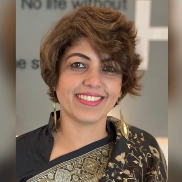 Smiling woman in a black and gold saree with short, styled hair, wearing long earrings, standing against a blurred background with text.