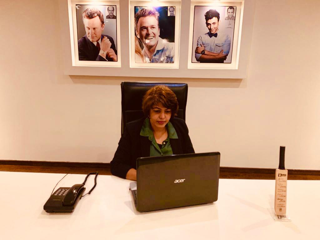 Woman in a black blazer seated at a white executive desk, working on a laptop in a modern office. Framed portraits of celebrities hang on the wall behind her.